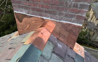 Newly installed copper flashing surrounds the base of a red brick chimney on a multicolored slate tile roof. The flashing is clean, reflective, and precisely bent to fit the contours of the chimney and roof valleys. Mortar seals the top edge of the flashing into the brick joints for waterproofing. The slate tiles show varying shades of green, gray, and purple, highlighting a high-end roofing installation.