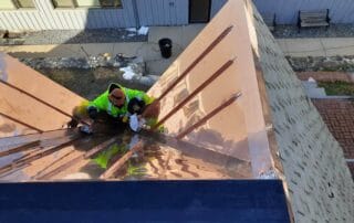 A worker in a high-visibility yellow jacket is crouched at the peak of a steep, newly installed copper roof, polishing or sealing the shiny surface. The roof features a complex multi-ridge design with raised seams for drainage. The bright, reflective copper contrasts with the building’s stone siding and modern architecture below. Benches, sidewalks, and landscaping are visible at ground level.