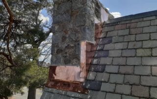 A stone chimney on a steep slate roof features freshly installed copper flashing along its base and vertical sides. The reflective copper contrasts sharply with the rough texture of the stone and the dark, weathered slate tiles. The work appears recently completed under a partly cloudy sky, with trees and a road visible in the background.