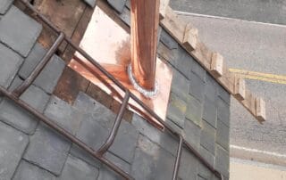 A steep slate roof in the process of having a copper vent pipe flashing installed. The bright copper base is fitted between weathered gray slate tiles, with a newly mounted cylindrical copper pipe extending upward. Wooden boards and a roof ladder are secured nearby for access. Below, a street and parked vehicles are visible, indicating an urban or suburban setting.