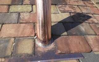A newly installed copper vent pipe protrudes from a multicolored slate roof. The pipe is fitted with a custom T-shaped copper flashing base that sits flush with the slate tiles. Behind it, a brick chimney with aged lead flashing is visible. The vibrant copper contrasts with the weathered reds, browns, and greens of the slate and the deep blue sky above.