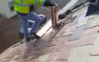 A worker in a high-visibility neon yellow jacket and jeans is installing copper flashing around a vent pipe on a steeply pitched slate roof. The multicolored slate tiles vary in shades of brown, red, and green. Tools and materials are scattered nearby, and safety ropes are anchored to the roof. The scene overlooks a residential neighborhood with houses, vehicles, and an American flag in the background.