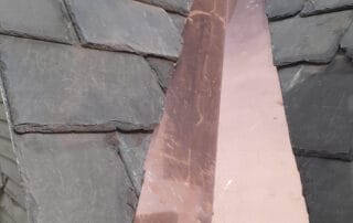 A close-up view of a copper valley flashing installed between two sections of a slate roof. The clean, reflective copper strip runs down the center, channeling water away from the roof valley. The surrounding dark slate tiles are irregular in shape and texture, emphasizing the traditional craftsmanship of the installation. The copper’s fresh appearance indicates recent work.