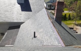 A high-angle view of a slate-tiled roof with a central valley, showing freshly installed copper flashing along the ridge and eaves. A red brick chimney and a white louvered cupola sit on separate roof sections. The surrounding neighborhood features residential homes and lush green trees under a clear blue sky, suggesting a sunny spring or summer day.