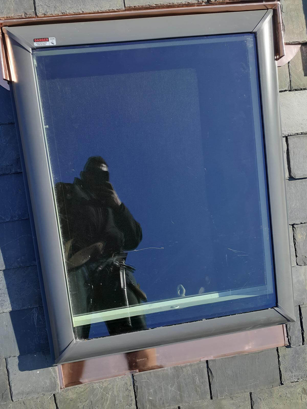 A close-up view of a modern skylight installed on a slate tile roof, surrounded by fresh copper flashing for waterproofing. The skylight’s glass reflects the image of the worker taking the photo. The copper flashing is neatly bent and secured beneath the slate tiles, enhancing both durability and aesthetics. A “Danger” sticker warning about tempered glass is visible in the top corner.