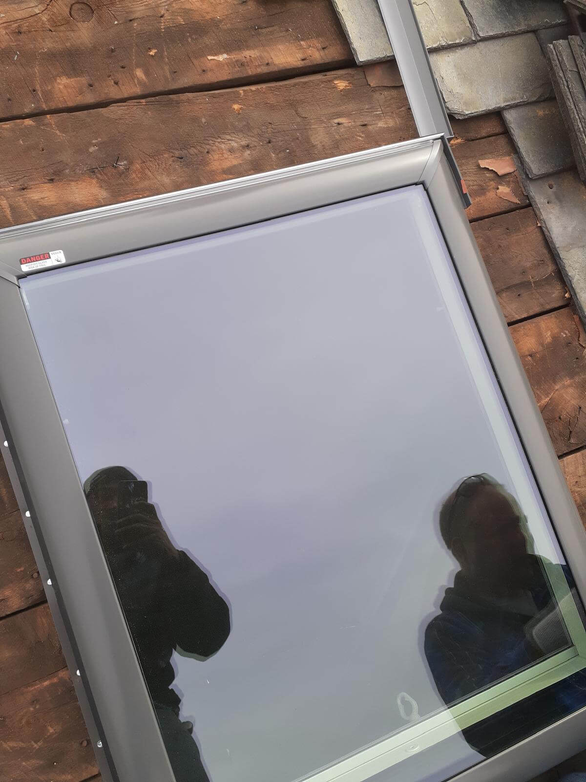 A skylight with a dark gray frame has been placed on an exposed wooden roof deck, awaiting final installation. The surrounding slate tiles have been temporarily removed. Two individuals are reflected in the skylight’s glass, one taking the photo and the other observing. A “Danger” sticker is visible in the top corner, warning of tempered glass.
