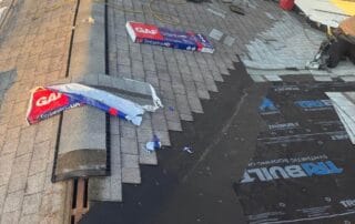 : A rooftop during active shingle installation, with asphalt shingles being laid over black TRI-BUILT synthetic underlayment. Roofing materials like GAF Timberline HD shingles, ridge caps, and nail guns are visible, along with tools and equipment. Snow covers parts of the surrounding yard, and two satellite dishes are mounted near the roof’s edge. The sun is low, casting a warm glow on the work in progress.