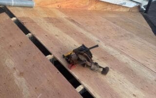 A partially completed roof deck with newly installed plywood sheets. A tool belt with various tools rests on the surface, and there are visible gaps where additional panels are yet to be installed. House wrap labeled “Lowe’s” and “HomeGuard Pro” is attached to the vertical wall, and snow is visible on the ground in the background. The scene is set during daylight near the edge of a wooded area.