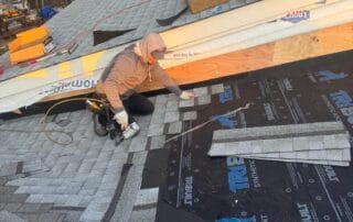 A roofer in a gray hoodie and gloves installs asphalt shingles over a black synthetic underlayment labeled “Tri-Built” on a sloped roof during sunset. Roofing materials, including shingles, insulation, and a nail gun, are spread around the work area. In the background, snow is visible on the ground and other rooftops, indicating winter conditions. The worker is mid-installation, carefully aligning the shingles for proper placement.