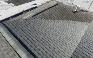 A roof with multiple intersecting slopes covered in light gray asphalt shingles, partially bordered by snow and ice buildup near the edge. A satellite dish and plumbing vent pipe are visible on the roof. The varying shades of shingles indicate a newer installation or drying patterns. Footprints in the snow and a partially exposed roof edge show ongoing maintenance or inspection during winter conditions.
