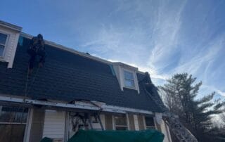 Two workers secured with harnesses are installing shingles on the steep slope of a residential roof with dormer windows. One is positioned on a rope while the other stands on a ladder. A green tarp covers part of the ground and the roof edge, and the sky is bright with wispy clouds and fading daylight. The house exterior features white siding and brown shutters.