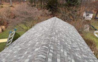 A symmetrical view of a gable roof covered in gray asphalt shingles with a prominent ridge vent cap along the peak. A ladder leans against the side, indicating recent or ongoing roofing work. The background features a grassy backyard with trees, bushes, a swing set, and a small white playhouse, suggesting a suburban residential setting in early spring or late fall.
