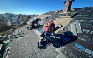 Two workers in safety harnesses are installing asphalt shingles on a pitched roof with black synthetic underlayment labeled “Tri-Built.” A skylight and chimney are visible on the roof, with shingles and tools scattered around. The background features residential buildings under a clear blue sky, indicating a sunny day during roof construction.