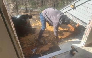A person in a hoodie and jeans is removing damaged material from a small roof deck area outside a house. The roof surface is heavily deteriorated with layers of decaying wood and black roofing felt. Tools, including drills, are on the ground nearby, and a large black trash bag filled with debris sits to the side. Trees and a road are visible in the background beyond the work area.
