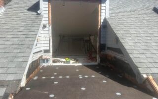 View from an open doorway onto a partially installed rooftop deck with exposed underlayment and fasteners. A can of roofing adhesive with a brush sits nearby, and a wooded area is visible in the background.