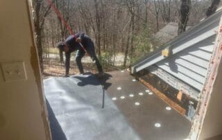 A worker is applying a black waterproof membrane over a flat roof deck using a roller tool. The underlayment with white fasteners has already been installed, and the exposed edge shows signs of prior water damage. The surrounding area includes a gray-sided house wall, visible interior framing, and a wooded yard in the background. Sunlight highlights the active progress of the roof repair.