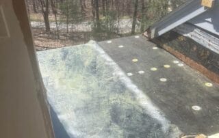 View from an open doorway onto a partially installed rooftop deck with exposed underlayment and fasteners. A can of roofing adhesive with a brush sits nearby, and a wooded area is visible in the background.