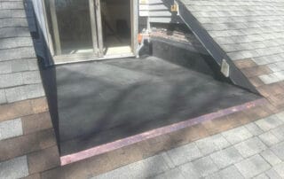 A small rooftop deck area with black rubber roofing, surrounded by sloped shingled surfaces and a sliding glass door leading into a house. Copper flashing is installed at the edge where the flat roof meets the sloped shingles.
