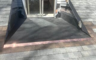 A small rooftop deck area with black rubber roofing, surrounded by sloped shingled surfaces and a sliding glass door leading into a house. Copper flashing is installed at the edge where the flat roof meets the sloped shingles.