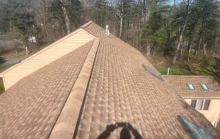 A symmetrical view down the ridge of a tan asphalt shingle roof, featuring multiple skylights and a clean ridge cap. The surrounding area includes a wooded backdrop and parts of nearby structures. The even pattern and fresh appearance of the shingles suggest recent installation. A person’s shadow taking the photo is visible in the foreground.