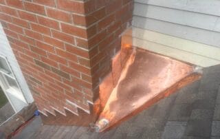 A close-up view of newly installed copper flashing around the base of a red brick chimney where it meets an asphalt shingle roof. The copper is clean and shiny, extending along the siding and roofline to ensure waterproofing. The installation includes step flashing on the chimney sides and a wide apron flashing along the bottom, forming a protective barrier against water infiltration.