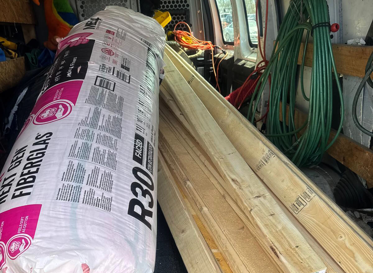 Construction Supplies Loaded in Work Van A van loaded with construction materials, including a large roll of R30 faced fiberglass insulation and several wooden boards, with green hoses and extension cords hanging on the side.