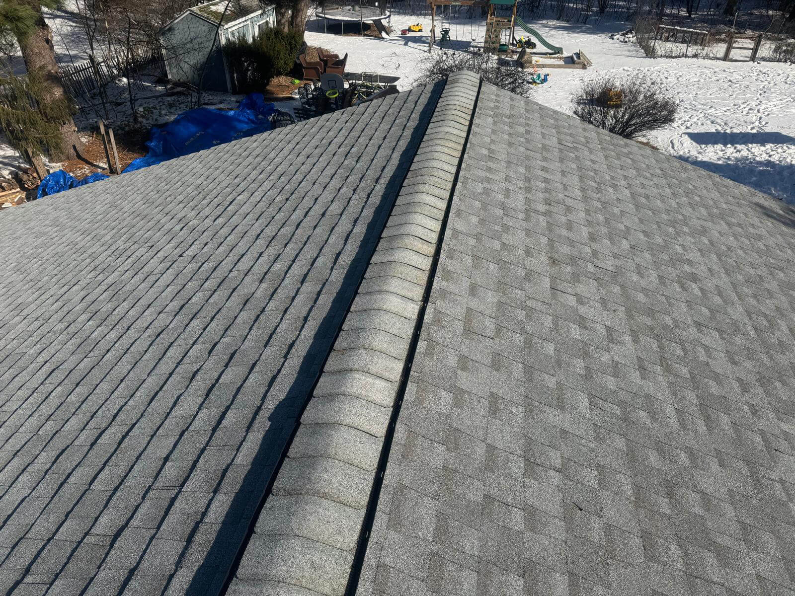 A well-aligned asphalt shingle roof viewed from the ridge, showcasing clean symmetry and uniform gray shingles. The central ridge vent runs along the peak, with neatly installed hip caps. Snow covers the surrounding yard, where outdoor items like a trampoline, chairs, and a children’s playground set are visible. The overall scene suggests recent roofing work completed during winter.