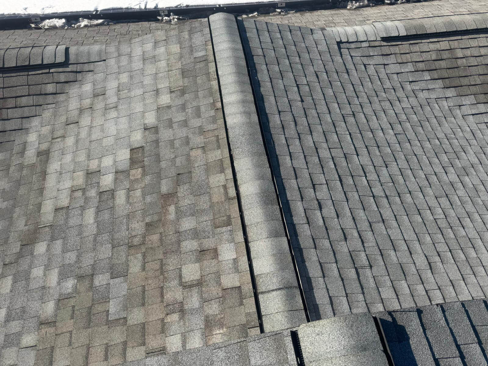 A close-up view of a complex asphalt shingle roof featuring intersecting slopes, a central ridge line with ridge vent caps, and well-aligned shingle patterns. The left slope shows a more varied shingle color blend compared to the right. Snow is visible along the roof’s edge, and the clean layout suggests recent installation.