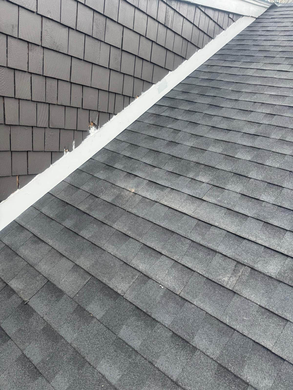 Close-up view of a residential roof showing gray asphalt shingles on the sloped surface and dark gray vertical siding shingles on the adjacent wall, with white flashing along the intersection.
