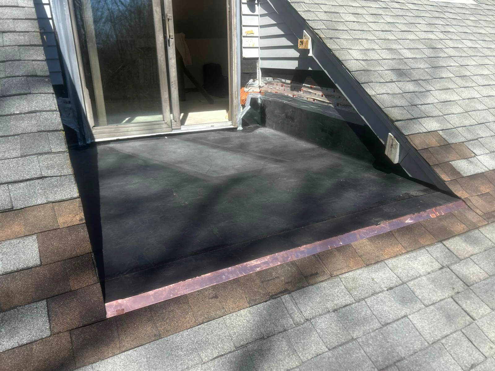 A newly finished flat roof section in front of a glass sliding door, surrounded by pitched asphalt-shingle roofing. The flat surface is sealed with a dark rubber membrane, and a strip of copper flashing lines the transition between the flat and sloped roof. The surrounding area shows clean lines and proper integration with the home’s siding and shingles, indicating a completed roof repair.