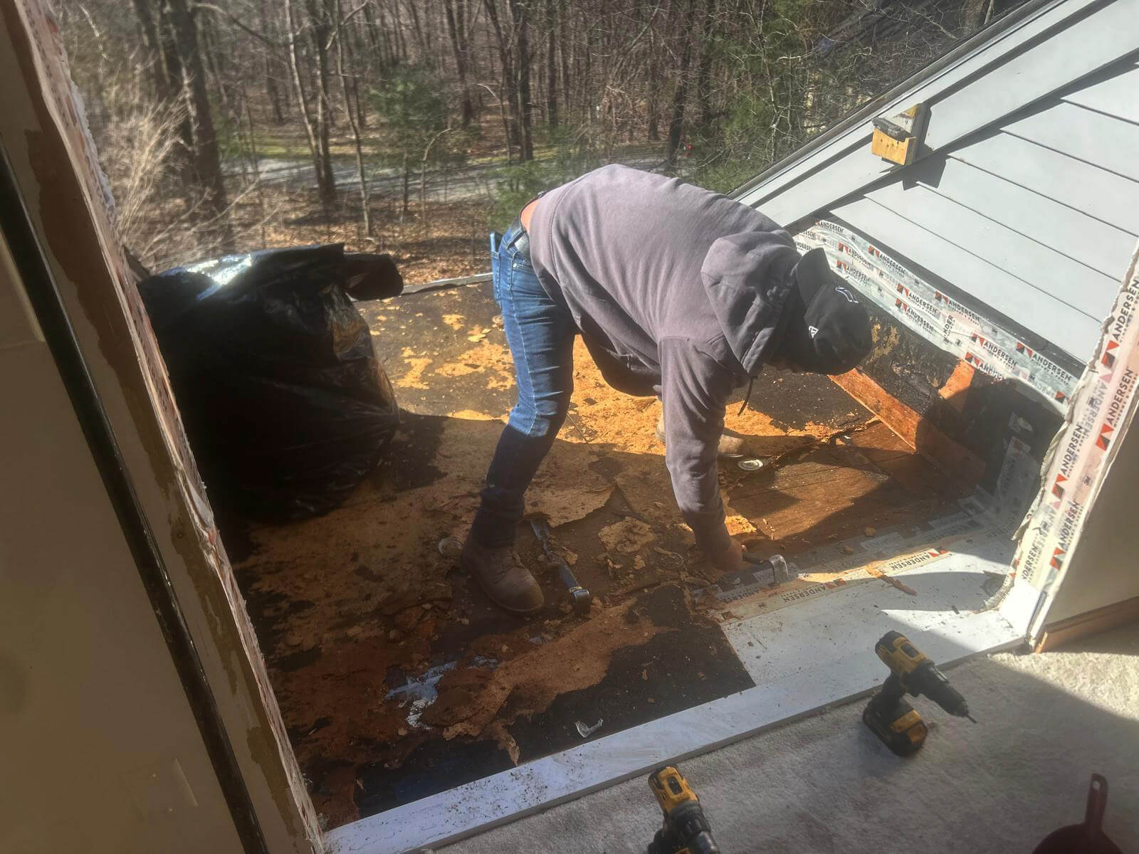 A worker wearing a hoodie, jeans, and boots is bent over removing damaged roof decking outside a home. The old material appears heavily rotted and is being stripped off. Tools such as drills are visible on the floor inside, and a large trash bag for debris sits to the left. The scene is lit by natural daylight with trees and a road visible in the background through the open roof area.