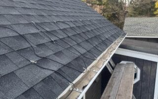 A close-up view of a roof with dark gray shingles, featuring heat cables installed in a zigzag pattern above the gutter, with a wooden plank scaffold and ladder setup for roof access.