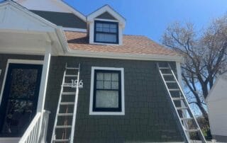 Two ladders leaning against the exterior of a green house with white trim and brown shingles, positioned under a second-story window and a roof dormer on a sunny day.