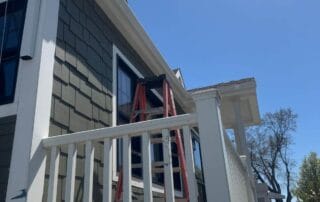 View of a white downspout and newly installed gutter system on a green-shingled house with white trim. A red ladder leans against the porch roof, and bright sunlight casts strong shadows on the deck and railing.