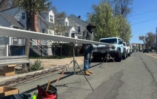 A worker stands beside long sections of white trim supported by tripods on a residential street, with tools and supply bags on the ground and white trucks parked nearby under clear blue skies.