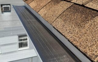 Detailed view of brown asphalt roof shingles and a newly installed metal gutter guard with a fine mesh screen, designed to prevent debris buildup, on a clear sunny day above a residential neighborhood.