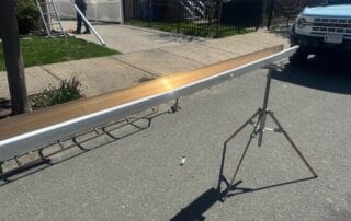 A long white gutter section resting on a metal stand in the street, with a worker in the background preparing a downspout and a parked Ford Bronco nearby.