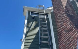 A tall extension ladder is propped up against a green-sided house with a brick chimney and white trim, reaching the roofline on a bright, clear day.