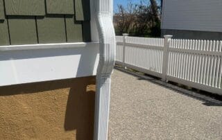 Close-up of a new white downspout with an offset elbow installed on the corner of a house with green siding and stucco base, next to a paved walkway and white picket fence, with a red reciprocating saw lying on the ground nearby.