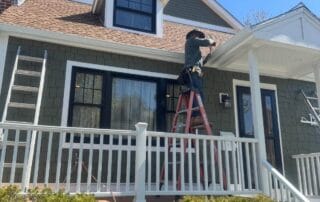 Worker standing on a ladder installing gutters on the front porch of a green Craftsman-style house with white trim, black window frames, and a brown shingle roof, under a clear blue sky.