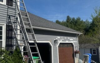 Gray house with a brown garage door, where patchwork spots are visible on the siding in preparation for painting. Multiple ladders are propped up against the wall, and painting supplies are scattered near the garage under a clear sky.