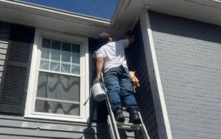 Man standing high on a ladder with a paint can, applying paint to the upper trim of a gray house with black shutters and white-framed windows under a clear blue sky.