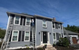 Wide view of a two-story gray house with black shutters and white trim, where a worker on a ladder paints near an upstairs window. Several ladders and drop cloths are set up around the house under a clear blue sky.