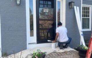 A man kneels while painting the white trim around a black front door of a gray brick house. Drop cloths cover the brick steps, and two flower pots with vibrant blooms sit on either side of the entryway.