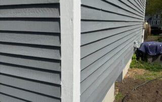 Close-up of a house corner showing newly painted gray horizontal siding with clean white trim and a concrete foundation, taken on a sunny day.