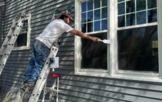 A construction worker standing on a ladder painting the white window trim of a gray-sided house, using a brush with a steady hand in bright daylight.