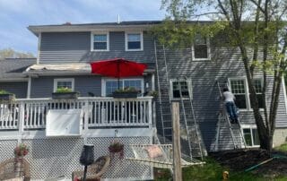 A residential two-story gray house with a white deck and red umbrella, where a worker on a ladder is painting window trim on the right side of the home surrounded by tall trees and yard tools.